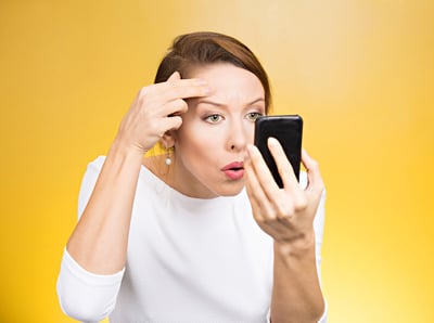 Closeup portrait, young, pretty beautiful woman surprised stunned to see zit on her face on pocket mirror, isolated yellow background. Negative emotion facial expression feelings, situation, reaction-1