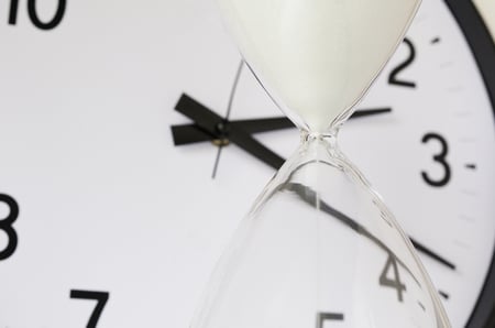 Two measurements of time White sand falling inside hourglass, with round analog clock in  background (focus on neck of hourglass), shallow depth of field